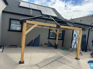 Wooden pergola structure with black roof panels installed over driveway next to residential home