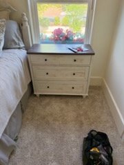 Bedroom corner with cream dresser, window with potted plant, and dog bed on carpeted floor