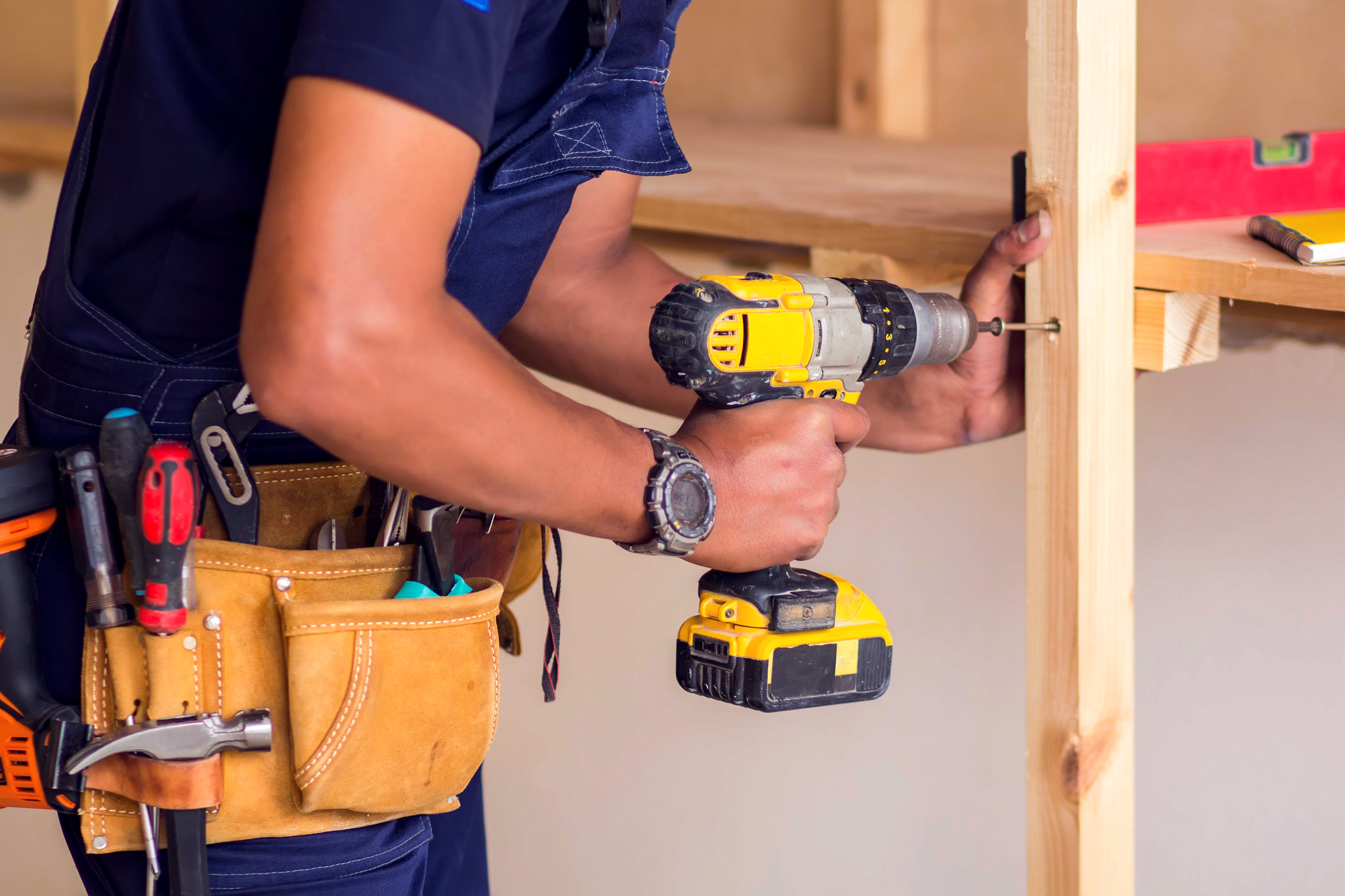 Construction worker using yellow cordless drill with tool belt on wooden frame