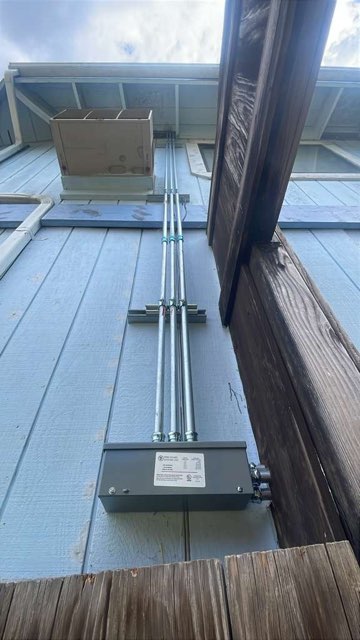 Metal conduit and electrical box mounted on blue metal roof panels between wooden beams